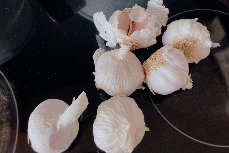 Several whole garlic bulbs, some with papery skins partially peeled, are scattered on a glossy black stovetop. One bulb reveals a glimpse of pinkish cloves inside.
