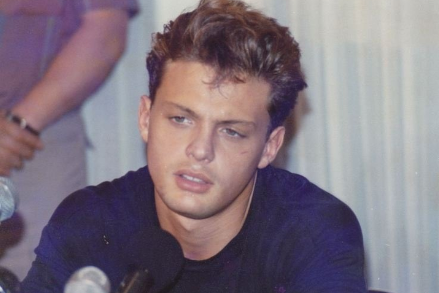 A young man with styled brown hair and a serious expression speaks at a press event, seated behind several microphones. He wears a dark shirt and is photographed indoors with a blurred figure in the background.