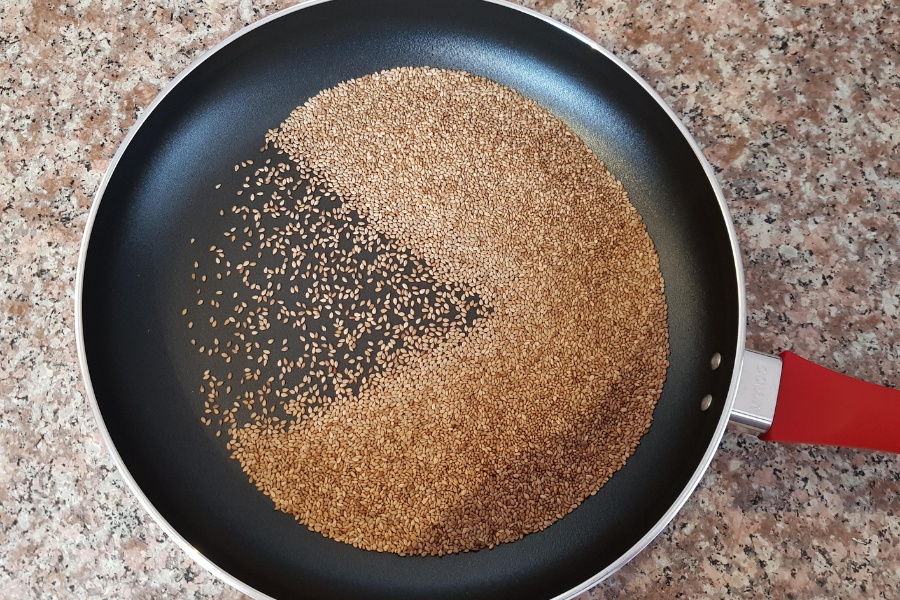Sesame seeds being dry-toasted in a non-stick frying pan, forming a distinct Pac-Man-like shape where some seeds are more toasted than others. The pan has a red handle and sits on a speckled kitchen countertop.