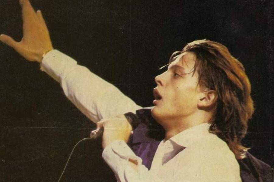 A young man with long, light brown hair is captured in profile, looking upwards with his mouth open as if singing. He is wearing a white collared shirt with the sleeves rolled up, and his left arm is raised high above his head, holding a microphone.