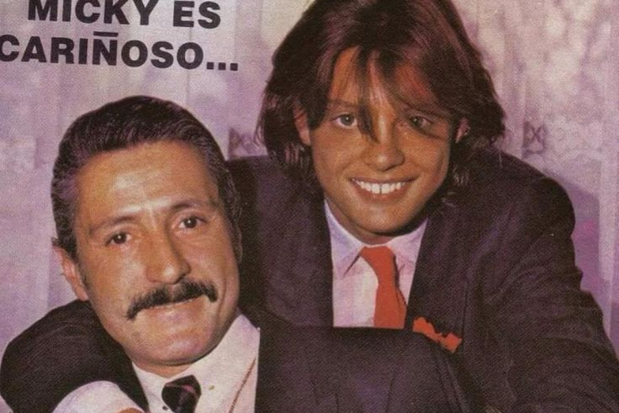 A man with a mustache and dark hair is in the foreground, wearing a suit and tie, smiling at the camera. Behind him, a younger man with long, dark hair and a red tie is embracing him from behind, also smiling broadly at the camera. Text in the background reads "MICKY ES CARIÑOSO...".