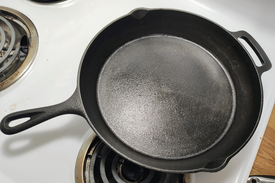 A seasoned cast iron skillet rests on the front burner of a white electric stovetop. The pan has a smooth, dark surface with a sheen from oil seasoning and features a long handle and helper handle.