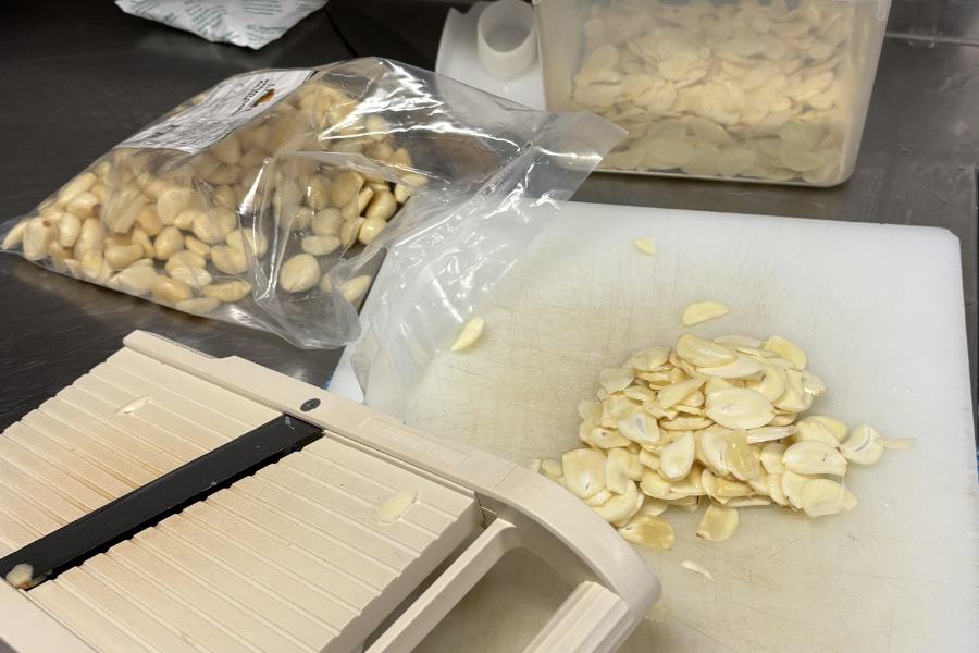 A pile of freshly sliced garlic sits on a white cutting board next to a mandoline slicer, with a bag of peeled garlic cloves and a container of sliced garlic in the background. The scene appears to be set in a commercial kitchen or prep area.