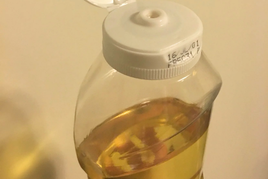 Close-up of a plastic squeeze bottle filled with golden-colored oil, likely vegetable or canola oil, with a flip-top cap. The expiration date is printed along the rim of the white cap.
