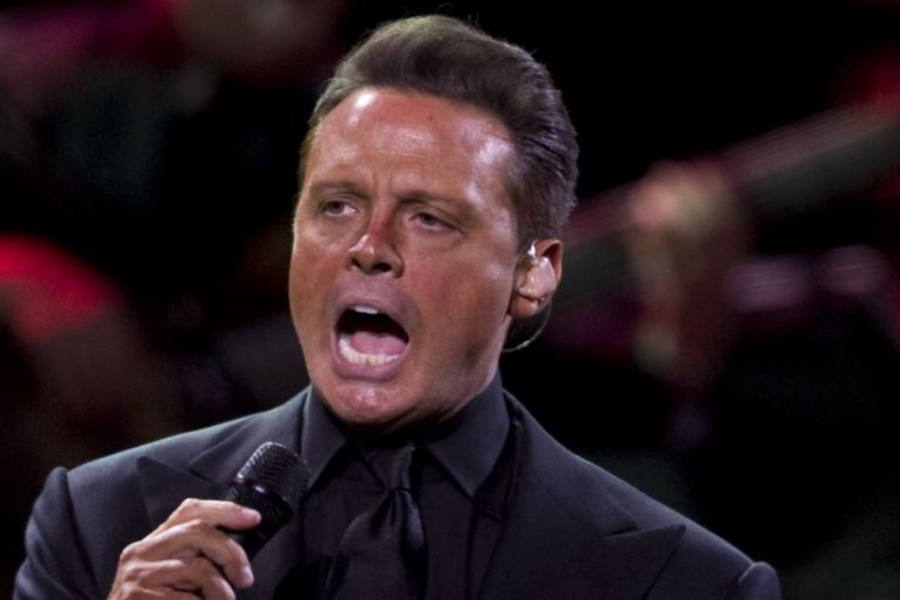 A man with dark, slicked-back hair and a tan complexion is captured mid-song, wearing a black suit and tie, holding a microphone. His mouth is wide open, and his eyes are looking upwards, suggesting intense vocalization.