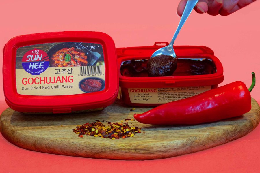 A tub of Sun Hee brand gochujang, labeled "Sun Dried Red Chilli Paste," is displayed on a wooden board alongside red chili flakes and a whole red chili pepper. A spoon scoops out the thick, dark red paste from the open container.