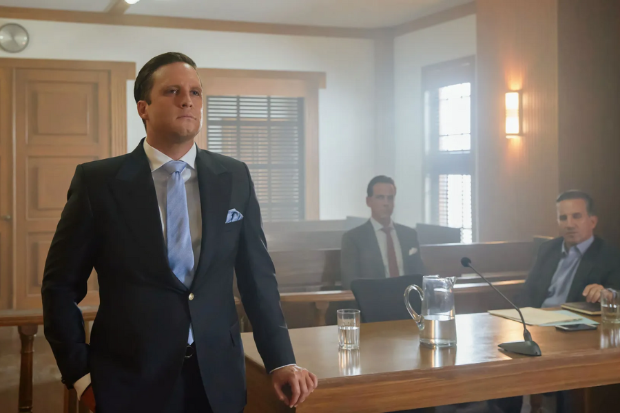A man in a dark suit, light blue tie, and white pocket square stands in a courtroom, looking seriously to the left. His left hand rests on a wooden railing. In the background, two other men in suits are seated at a table with a pitcher of water and a microphone.