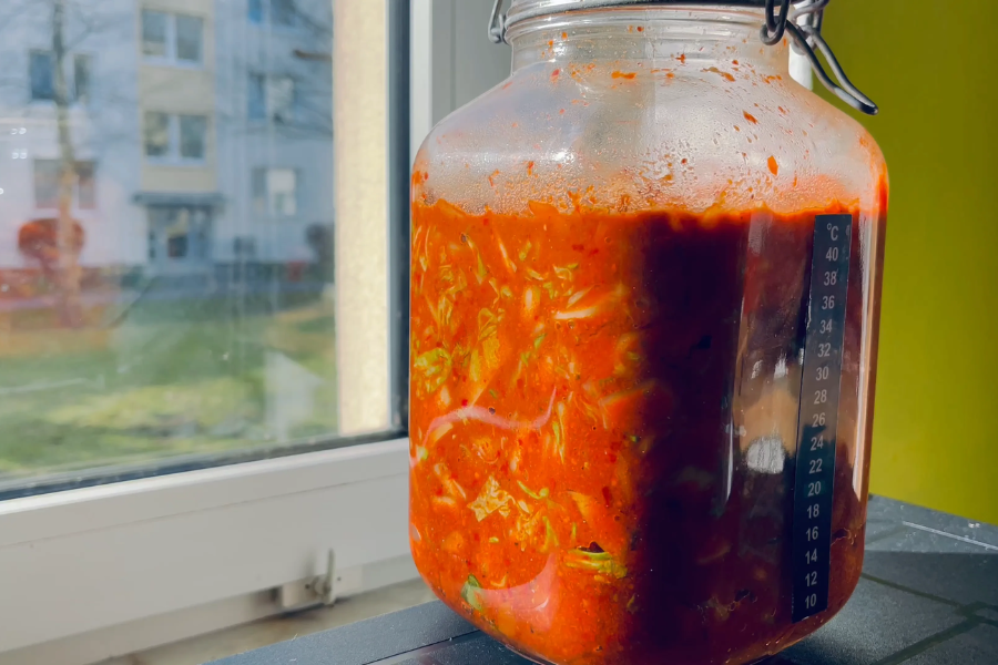 A large glass jar filled with bright red homemade kimchi sits on a windowsill, bathed in sunlight. Visible through the jar are shredded cabbage, red chili paste, and other fermenting vegetables, with a temperature strip on the side showing fermentation conditions.