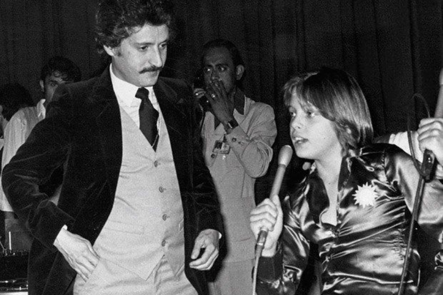 Black-and-white photo of a young boy in a shiny button-down shirt holding a microphone, speaking animatedly to a man in a three-piece suit with a mustache. The scene appears to be a public event or interview, with other people and microphones in the background.