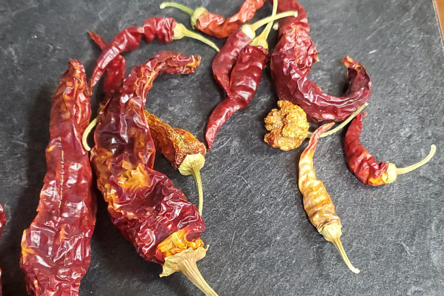 A variety of dried red chili peppers, some long and wrinkled and others short and round, are scattered across a dark stone surface. The peppers range in shades from deep red to reddish-orange with some yellowing tips.