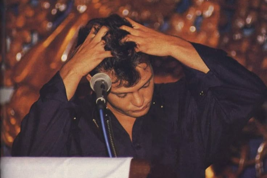 A man with dark hair, wearing a dark shirt, stands with his hands running through his hair. He is looking down and appears to be in thought or distressed. A microphone is in front of him.