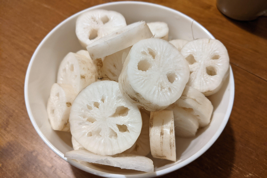 A white bowl filled with thick slices of peeled lotus root, each piece showing the vegetable's distinctive hole pattern. The slices are slightly off-white and laid unevenly on top of each other.