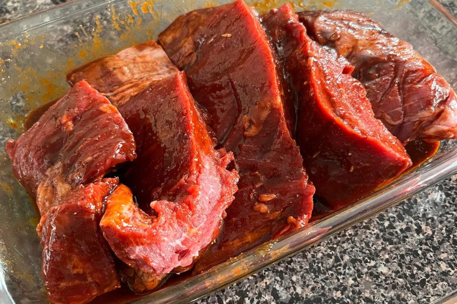 Thick cuts of raw beef are coated in a red marinade and arranged in a glass container, ready for cooking. The meat has a glossy texture from the marinade, which appears to include spices and possibly soy or chili paste.