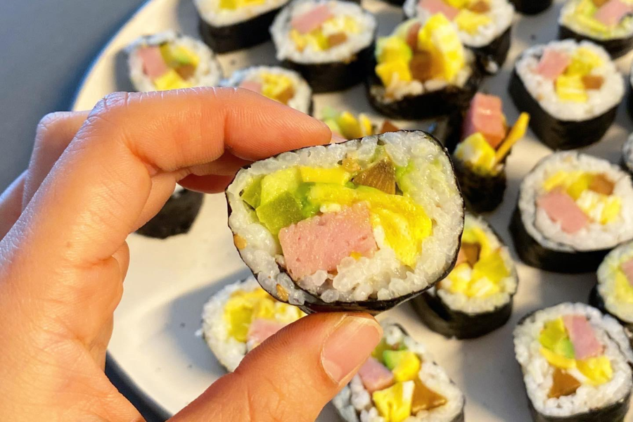 A hand holds up a piece of kimbap, showing its colorful filling of rice, yellow egg, pink ham, green avocado, and pickled vegetables wrapped in seaweed. A plate full of similar sliced kimbap pieces is visible in the background.