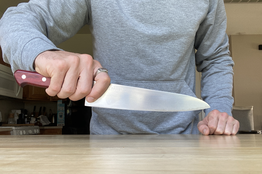A person in a gray hoodie stands behind a wooden counter holding a large kitchen knife with a wooden handle, blade pointing downward in a firm grip. The kitchen setting is visible in the background.