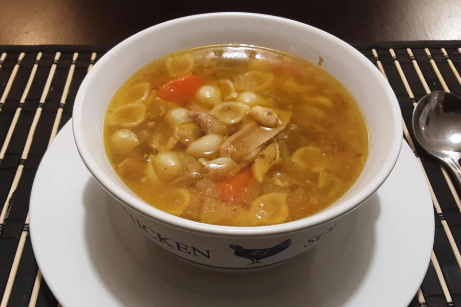 A bowl of homemade chicken soup filled with small pasta shells, shredded chicken, carrots, and pearl onions sits on a white plate atop a bamboo placemat. A spoon rests to the side of the bowl.