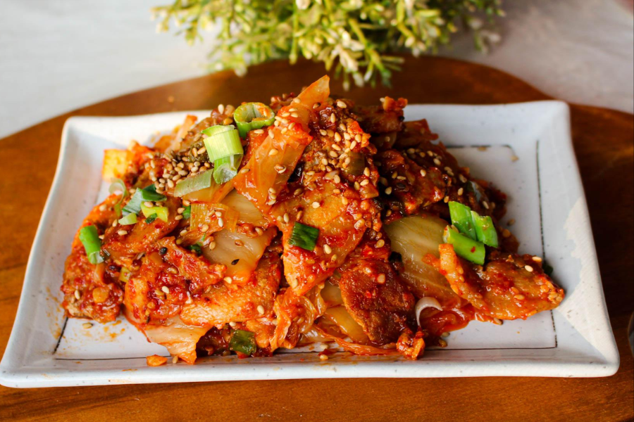 A serving of stir-fried kimchi is presented on a rectangular white plate, garnished with sesame seeds and chopped green onions. The vibrant red hue of the fermented cabbage is coated with chili paste and oil.