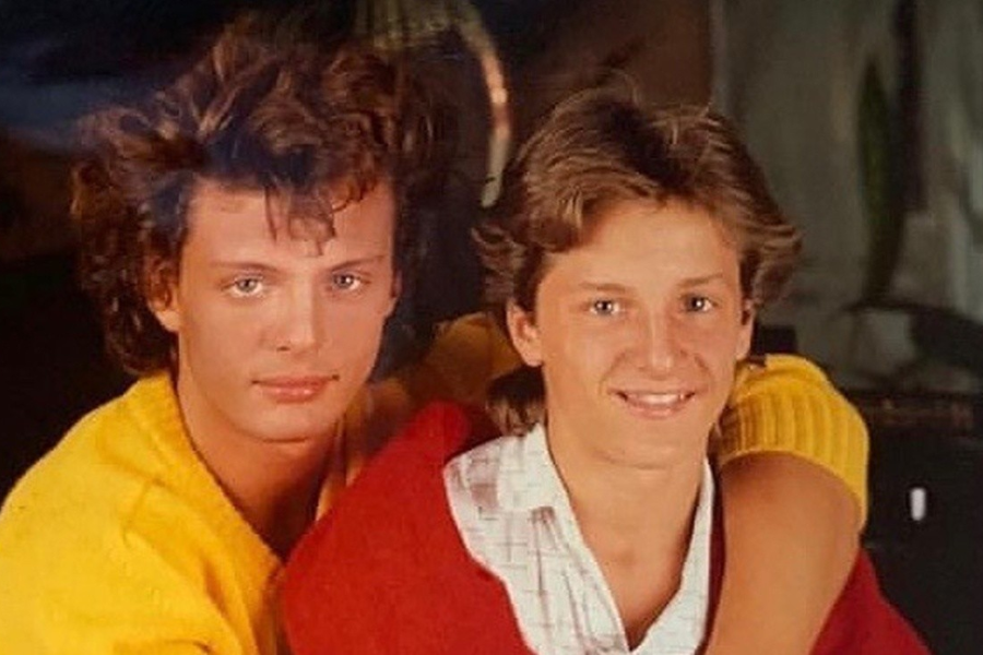 Two young men with similar light brown, wavy hair are positioned closely together. The man on the left wears a yellow top and looks directly at the camera with a serious expression. The man on the right, smiling widely, wears a plaid shirt under a red and yellow garment, with his arm around the other man.