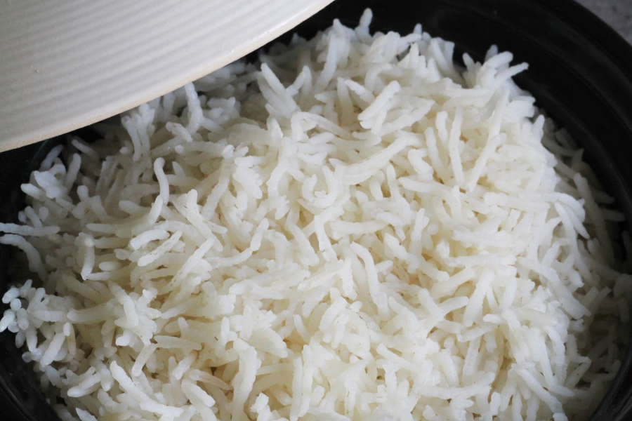 A pot of freshly cooked white rice with long, fluffy grains is partially uncovered by a white lid. The rice appears soft and perfectly steamed, ready to serve.
