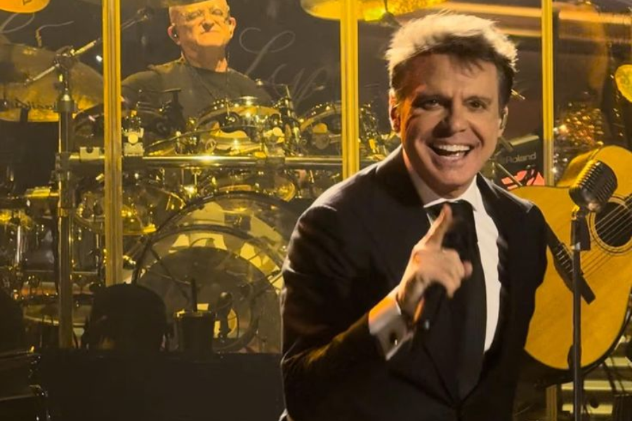 Luis Miguel, a man with light brown, slicked-back hair, is on stage in a black suit, white shirt, and black tie, smiling widely and pointing his finger. In the background, a drummer is visible behind a gold drum kit. A guitar and microphone stand are also on stage.