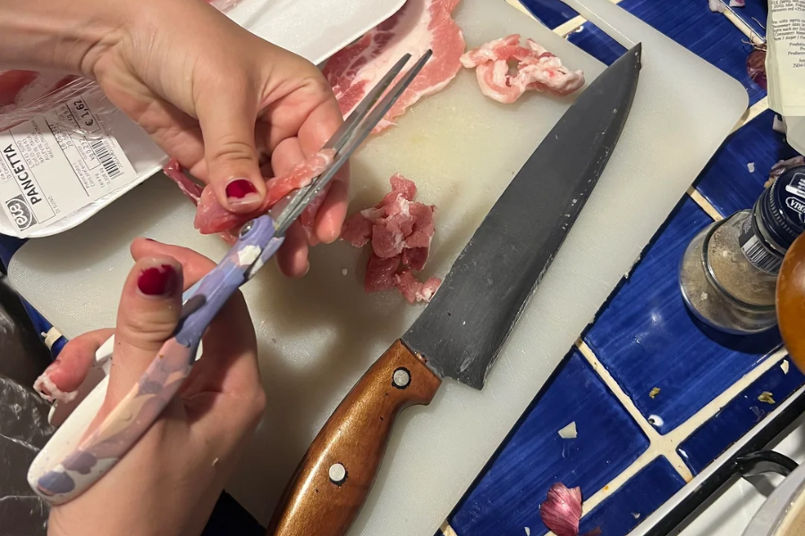 A person with red nail polish uses kitchen scissors to cut raw pancetta over a white cutting board. A chef’s knife, pepper grinder, and food packaging labeled “Pancetta” are nearby on a blue-tiled countertop.