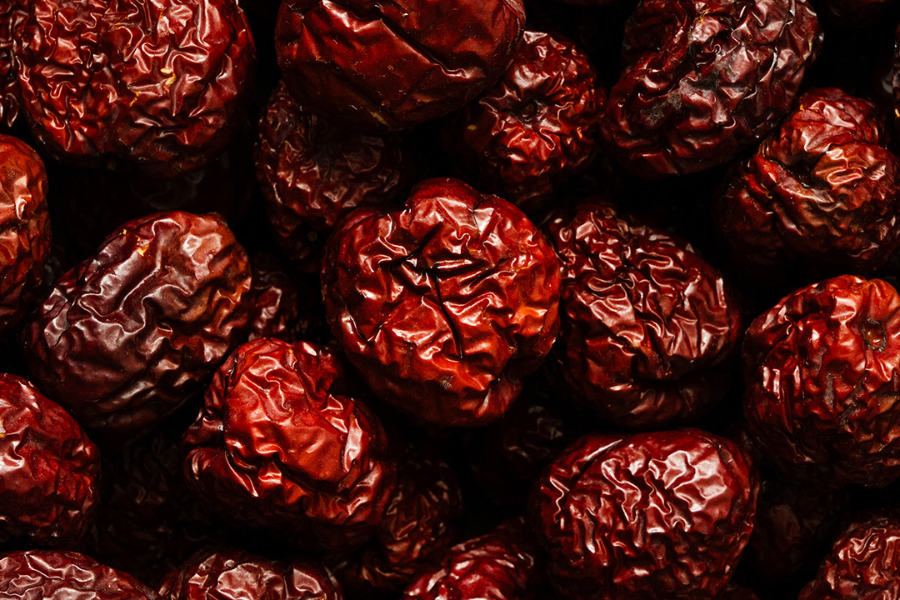 A close-up of a pile of dried red jujubes, also known as red dates, with their signature wrinkled skin and deep reddish-brown color. The texture is rough and slightly glossy, indicating their chewy consistency.