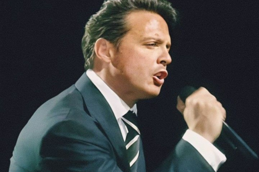 A man with dark, slicked-back hair is captured in profile while singing into a microphone. He is wearing a dark suit, a white shirt, and a striped tie, with his mouth open in mid-song.
