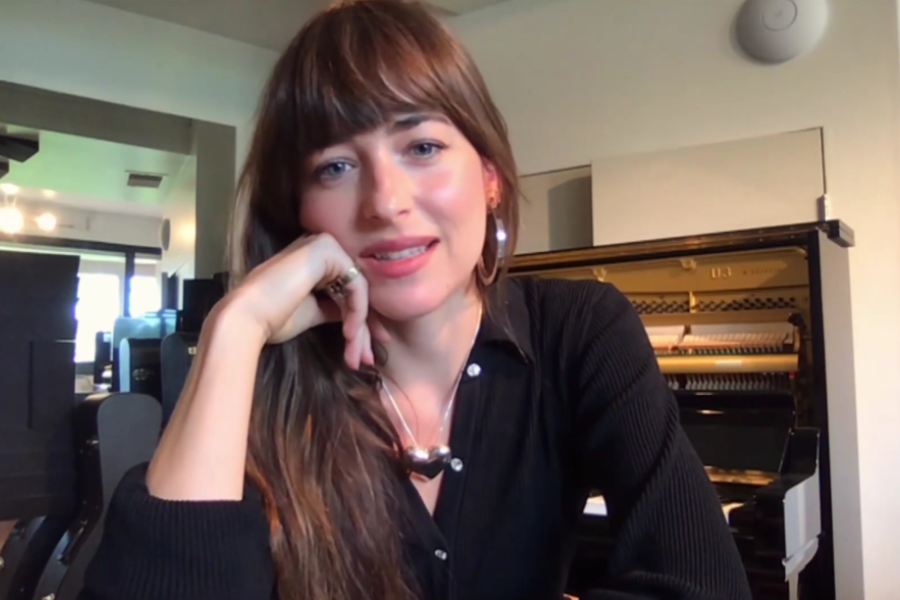A woman with long brown hair and bangs, wearing a black top and hoop earrings, sits in front of an upright piano in a modern room, resting her chin on her hand and smiling slightly toward the camera.