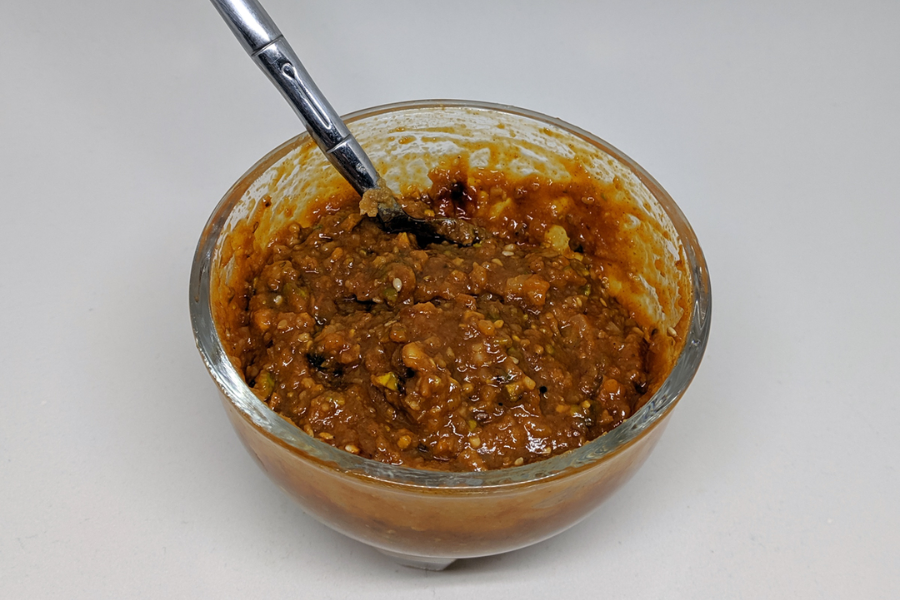 A glass bowl filled with a chunky, reddish-brown chili paste or sauce, with visible seeds and flecks of ingredients, sits on a white surface. A spoon is partially submerged in the thick mixture.