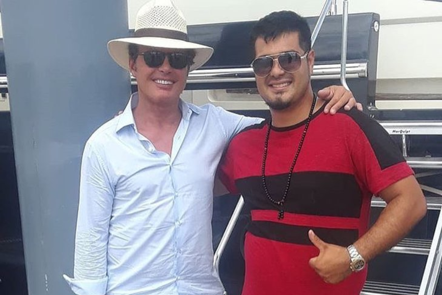 Two men are standing outdoors, likely on a boat or by the water. The man on the left wears a light-colored fedora hat, sunglasses, and a light blue collared shirt, smiling at the camera. The man on the right wears sunglasses, a red and black striped t-shirt, and a long black beaded necklace, with his arm around the other man's shoulder and his thumb up.