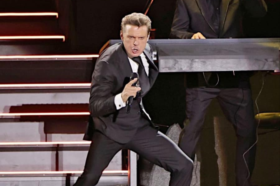 A man with slicked-back hair, wearing a black suit, white shirt, and dark tie, is singing passionately into a microphone on stage. He is leaning forward with one leg bent, and his mouth is open wide. Behind him to the right, another person is partially visible behind a keyboard or soundboard. There are illuminated steps to the left.