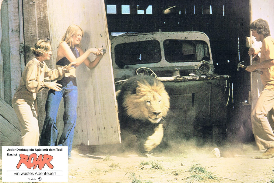 A lion bursts out of a dusty garage or barn as three people—two women and one man—react in alarm, one woman holding a camera while the man grips a hammer; the chaotic scene is part of a promotional image for the film Roar, with German text in the corner translating to “Every shoot a game with death!”