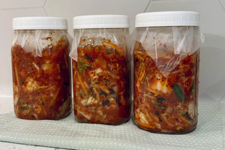 Three jars of homemade kimchi packed with napa cabbage, green onions, and spicy red pepper paste sit on a light cloth surface. The jars are sealed with white lids and lined with a layer of plastic wrap under each cap for extra fermentation protection.