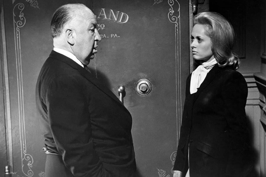 Director Alfred Hitchcock faces actress Tippi Hedren in a tense black-and-white scene, both dressed formally and standing in front of an ornate vault door labeled “Sutherland” in a dramatic setting suggestive of a film noir moment.