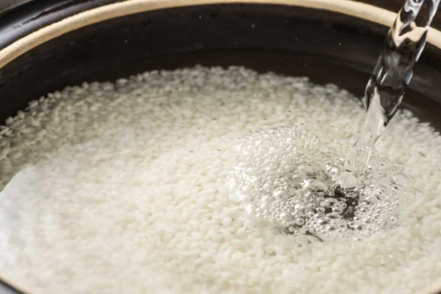 Close-up of water being poured into a pot filled with uncooked short-grain rice, creating bubbles as it hits the surface. The rice appears to be in the soaking stage before cooking.