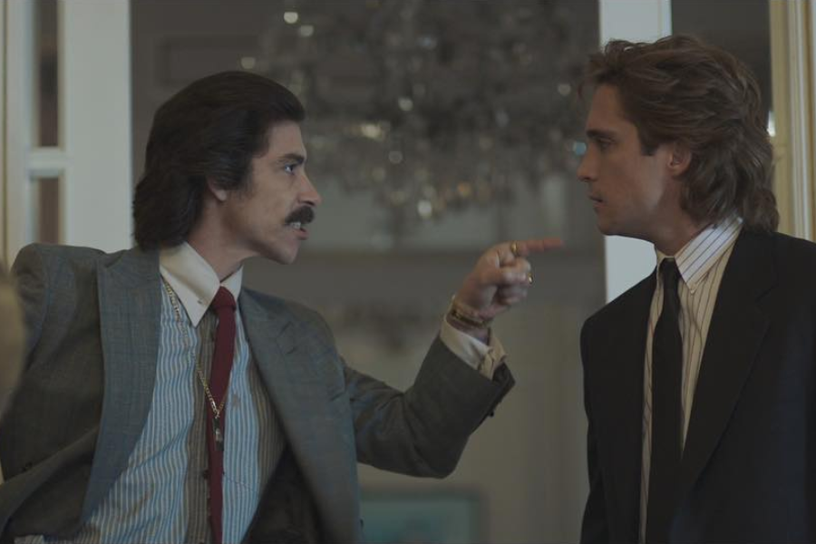 Two sharply dressed men face off in a tense confrontation indoors. The man on the left, with a mustache and slicked-back hair, points his finger angrily at the other man, who looks back at him calmly, both wearing suits and ties under dramatic lighting.