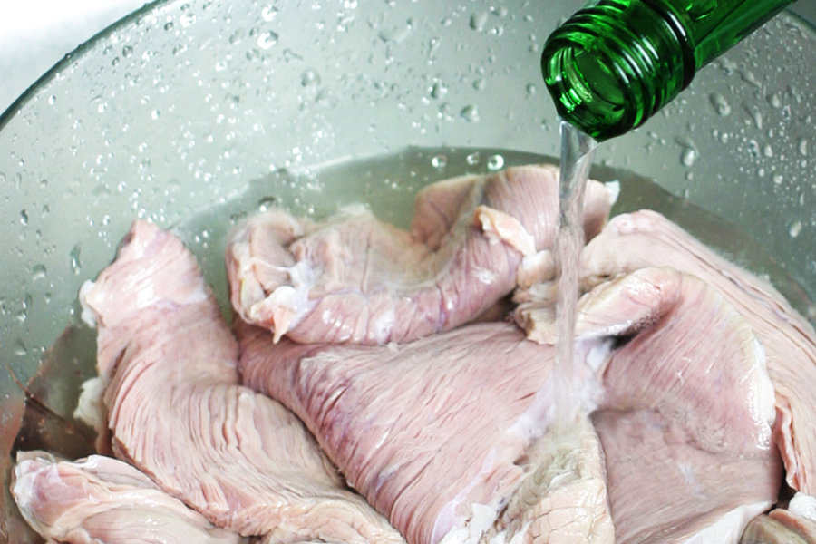A close-up of raw beef tripe soaking in a large bowl of water, while clear liquid—likely vinegar—is being poured in from a green bottle for cleaning. Water droplets are visible on the sides of the bowl.
