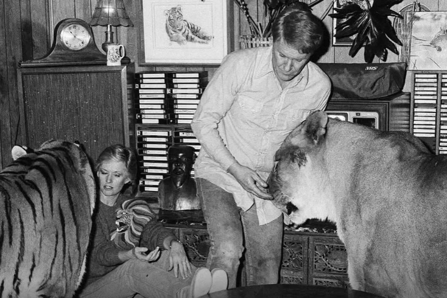 In a vintage black-and-white photo, Tippi Hedren sits on the floor beside a tiger while a man, likely Noel Marshall, stands next to her feeding a lion in a wood-paneled room filled with VHS tapes, art, and eclectic decor, blending domestic life with exotic animals.