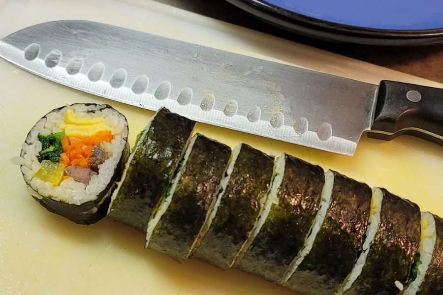 A sliced roll of kimbap sits on a cutting board beside a chef’s knife, with visible colorful fillings like carrot, spinach, egg, and pickled radish wrapped in seaweed and rice. One piece is angled to display the vibrant ingredients inside.