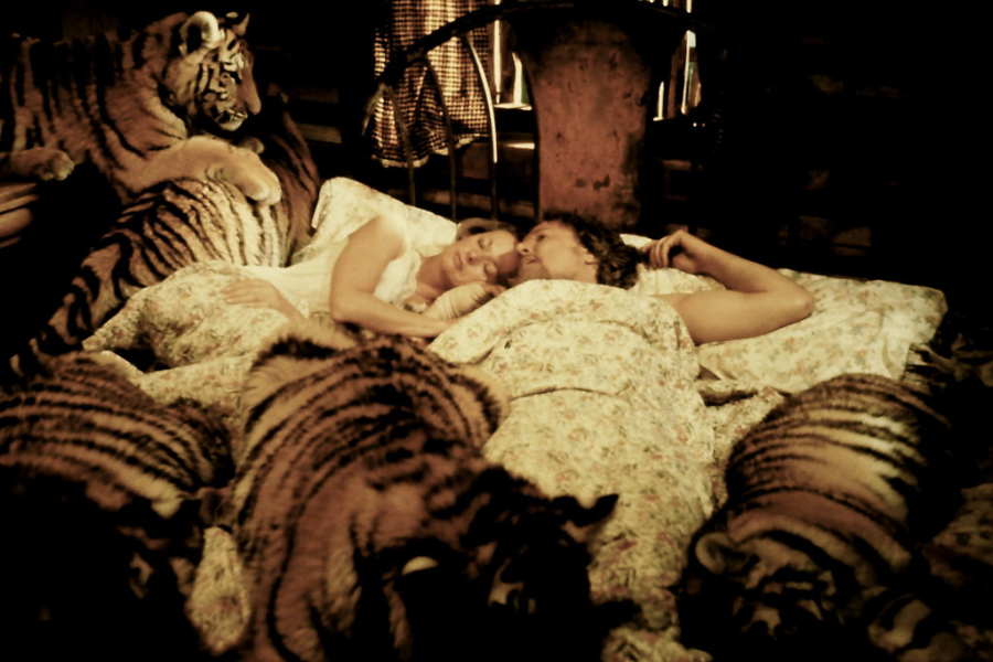Two people lie in bed under floral sheets, resting closely together, surrounded by several tiger cubs—some on the bed and others perched beside it—inside a dimly lit, rustic cabin bedroom.