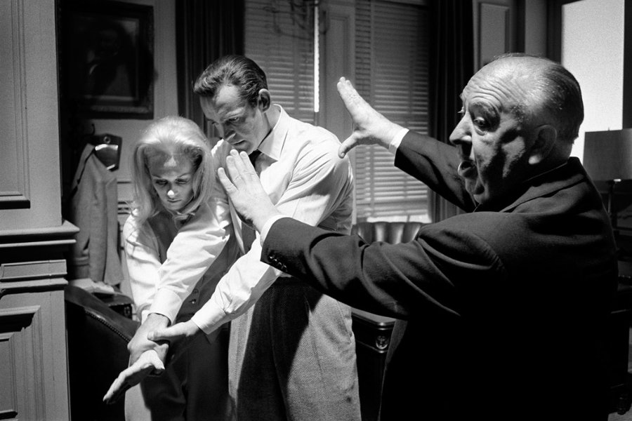 Alfred Hitchcock directs Tippi Hedren and a male actor on set, dramatically demonstrating a gesture with raised arms as the two actors closely follow his lead in a dimly lit office-like setting, captured in striking black and white.
