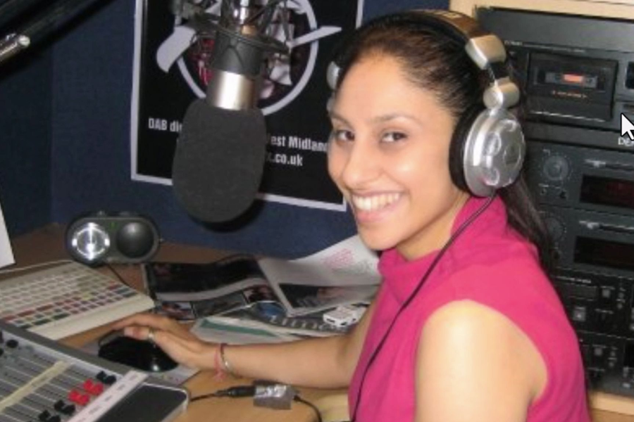 A smiling woman wearing large over-ear headphones sits at a radio broadcasting station, surrounded by sound equipment, a mixing board, and a large microphone. She is wearing a sleeveless magenta top and looking at the camera while using a computer mouse.