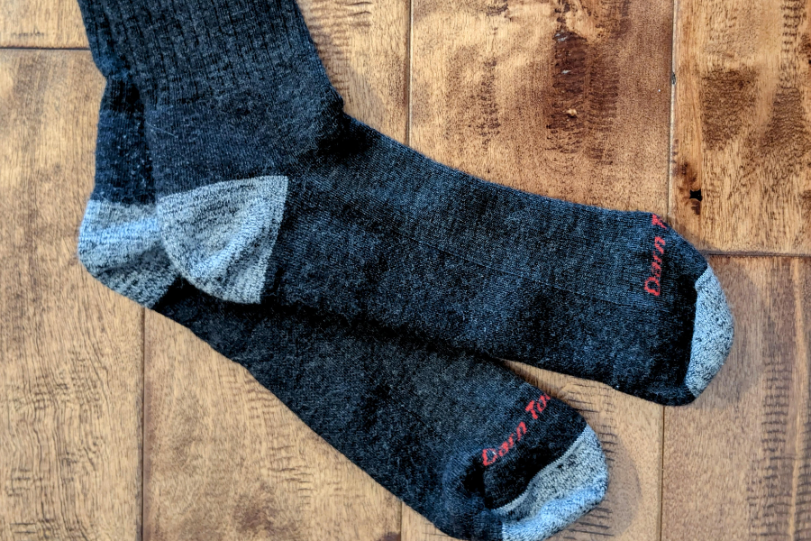 A pair of dark gray wool socks with light gray heel and toe accents, laid out on a wooden floor. The toe area of each sock is embroidered with red text that reads "Darn Tough."