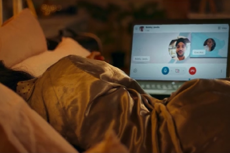 A woman lies in bed wearing a silky robe, facing a laptop screen displaying a video call. On the screen, two participants are visible: Bobby Jandu and someone named Cree Ann, suggesting a group video chat in progress.