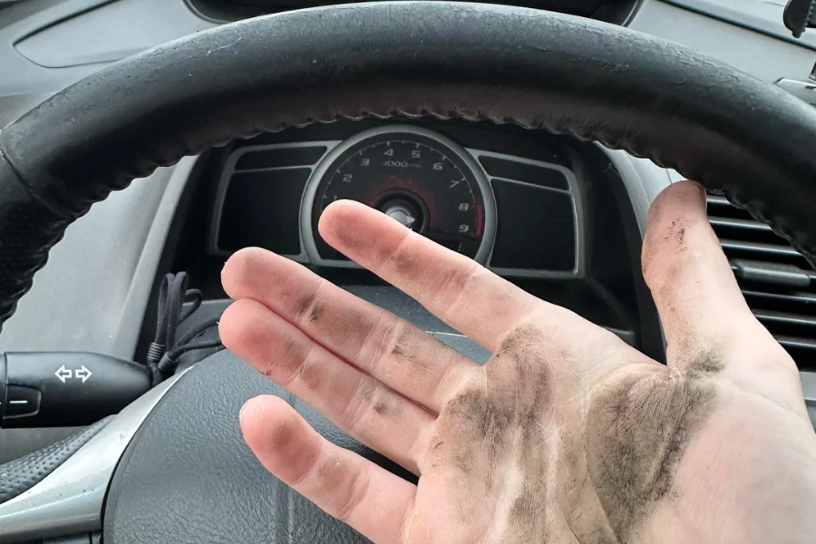 A person's hand covered in black grease or dirt is held up in front of a car steering wheel, with the dashboard and speedometer visible in the background.