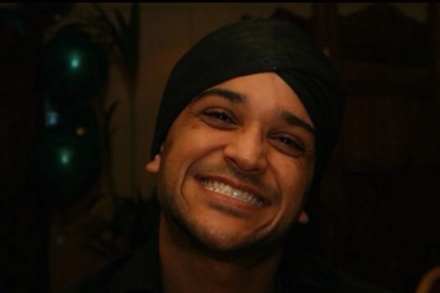 A man wearing a black turban and dark shirt smiles widely at the camera in a warmly lit indoor setting, with green balloons and plants visible in the background.