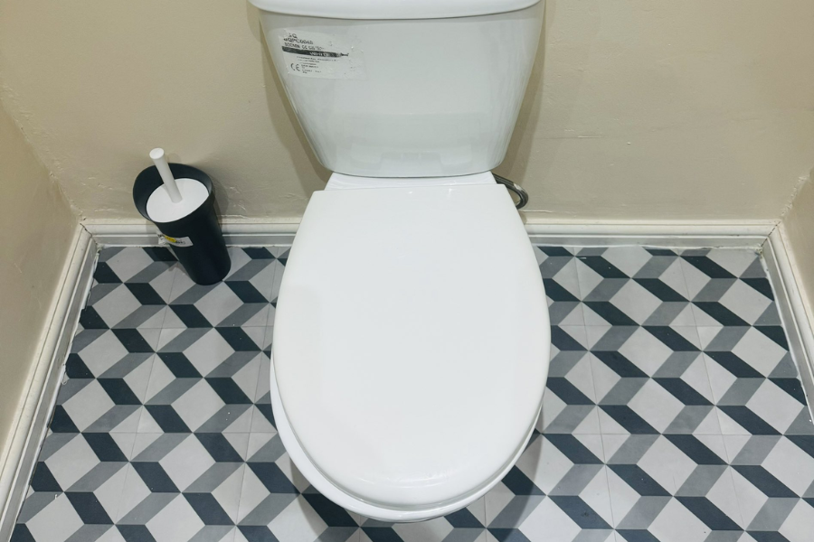 A white toilet with the lid closed, positioned against a beige wall and placed on a patterned black, white, and gray geometric tile floor. A toilet brush in a black holder is placed to the left of the toilet.