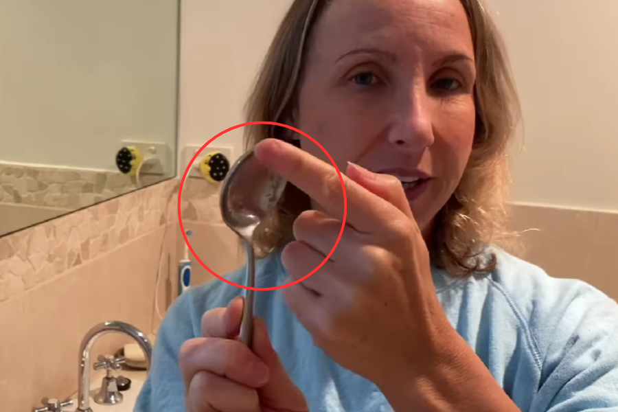 A woman in a blue shirt stands in a bathroom, pointing to the back of a metal spoon she is holding upright, with a red circle digitally highlighting the area she's referencing.