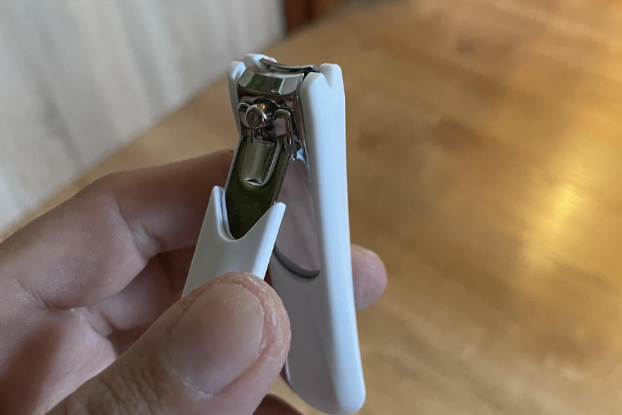 A hand holding a silver nail clipper with a white plastic grip, positioned over a wooden floor in a well-lit room.