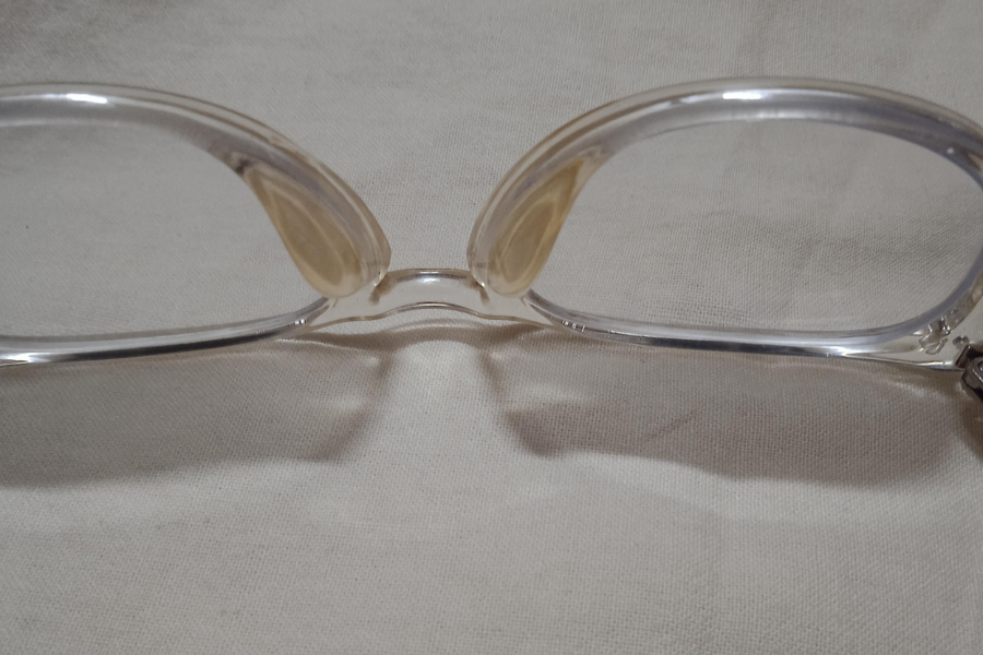 Close-up of a pair of clear-framed eyeglasses resting on a light fabric surface, showing visible yellowing on the inner nose pads.